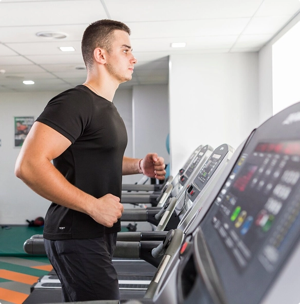 A man running on a treadmill in a gym, focused on his workout amidst various gym equipment in the background.