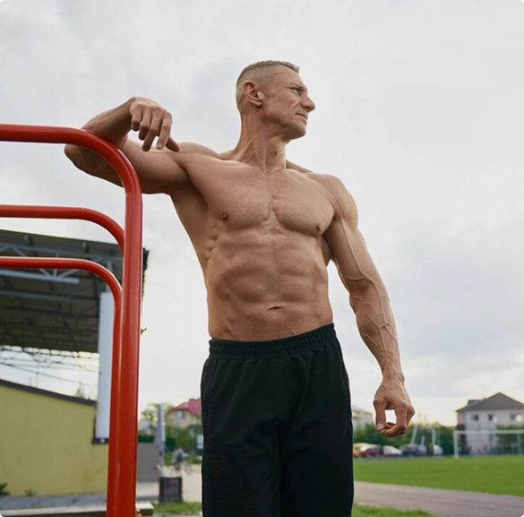 A shirtless man balances on a red rail, exuding a casual demeanor in a simple outdoor setting.