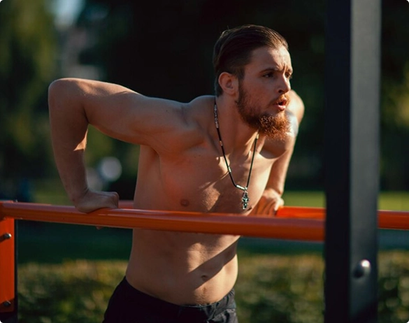 A shirtless man performs pull-ups on an orange bar, showcasing strength and fitness in an outdoor setting